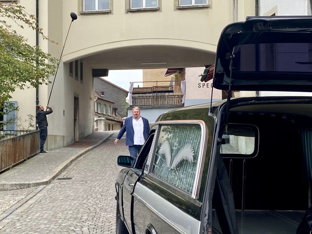Mike Müller läuft mit einem Koffer in Richtung des Leichenwagens.